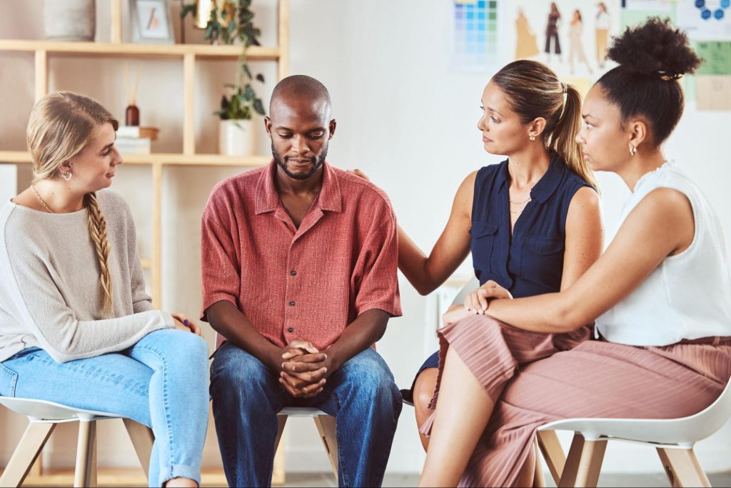 Support group sitting in a circle with a shelf in the background.