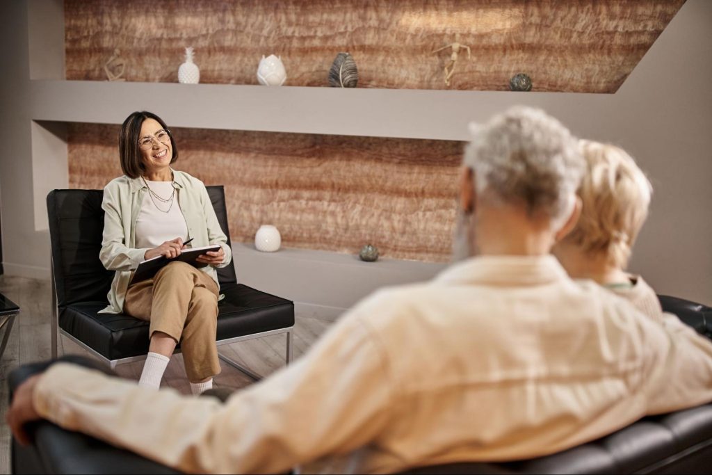 A female therapist with glasses smiling and holding a notepad across from a married couple in a therapy session.